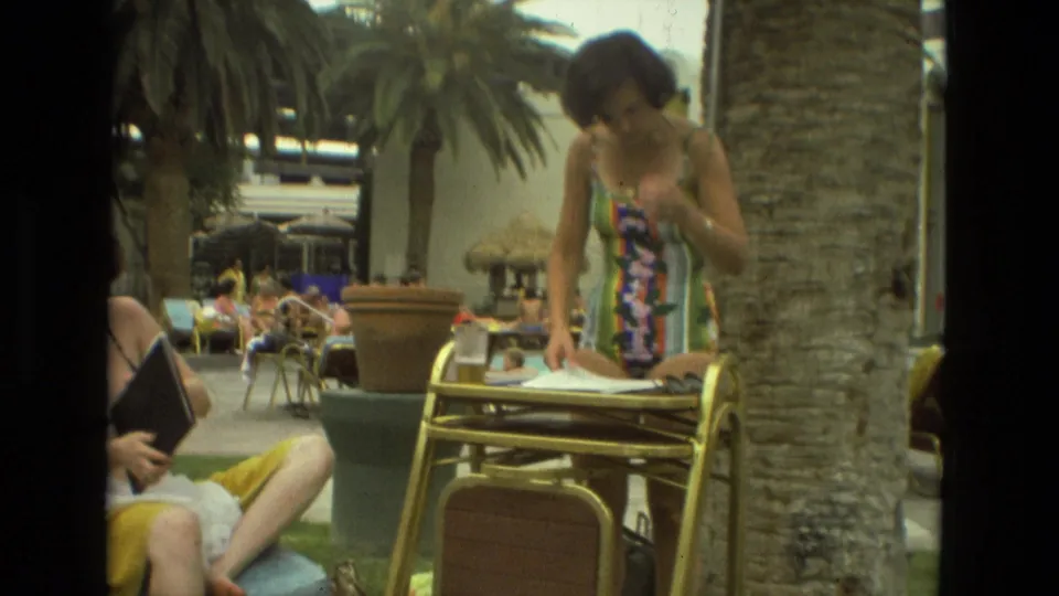Woman in a colorful swimsuit organizes papers on a gold metal patio table at a 1978 Las Vegas resort.