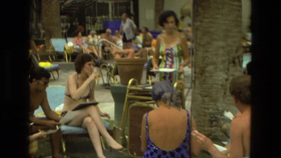 At a 1978 Las Vegas pool area, a woman in a bright striped dress stands holding papers near seated guests.