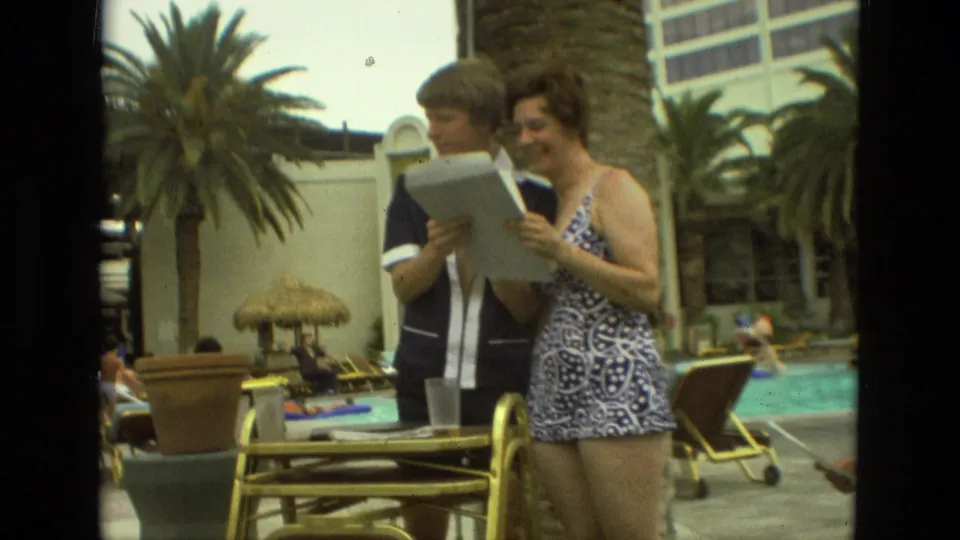 1978 Las Vegas resort-pool scene showing two adults reading a paper together with palms, chairs, and a thatched umbrella nearby.