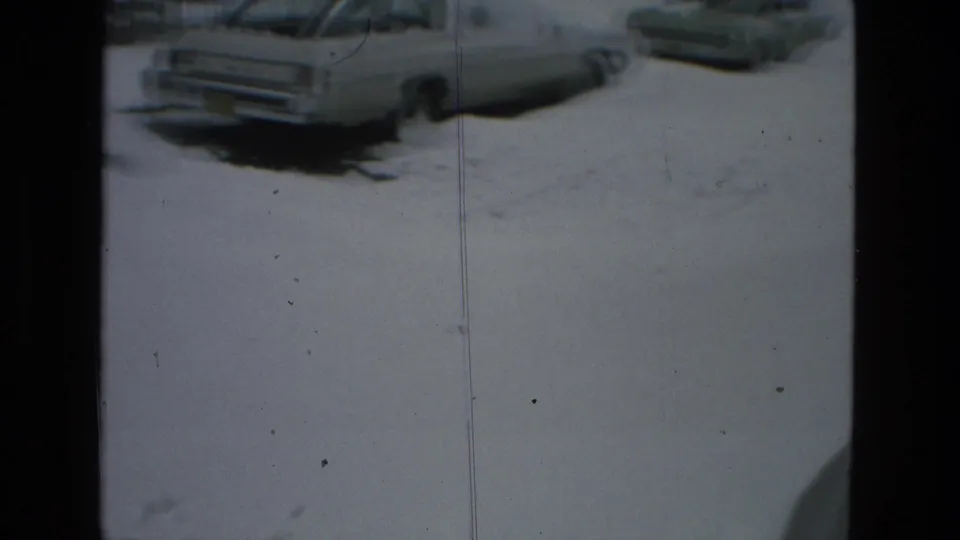 1978 Drexel Hill home-movie frame showing a light-colored car parked along a snow-covered residential street, with another vehicle partly visible nearby.