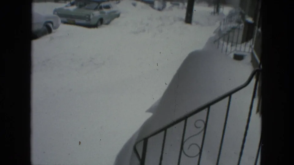 1978 Drexel Hill 8mm frame from an elevated stoop or porch, showing a wrought-iron railing, deep snow, and parked cars along the residential street.