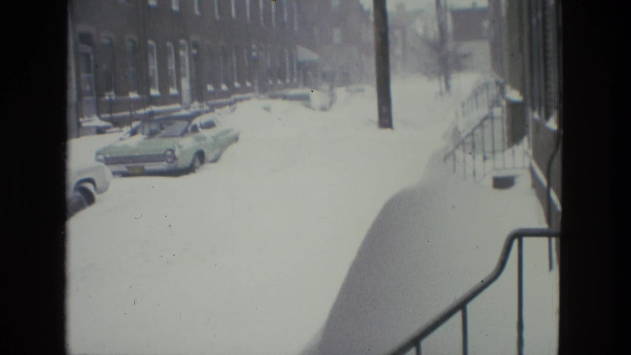Snow-covered Drexel Hill street seen through a home-movie frame, with parked cars, brick rowhouses, utility poles, and heavy sidewalk snow.