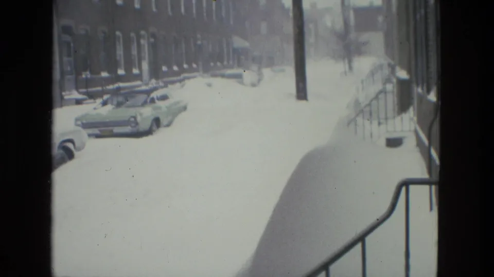 Snow-covered Drexel Hill street seen through a home-movie frame, with parked cars, brick rowhouses, utility poles, and heavy sidewalk snow.