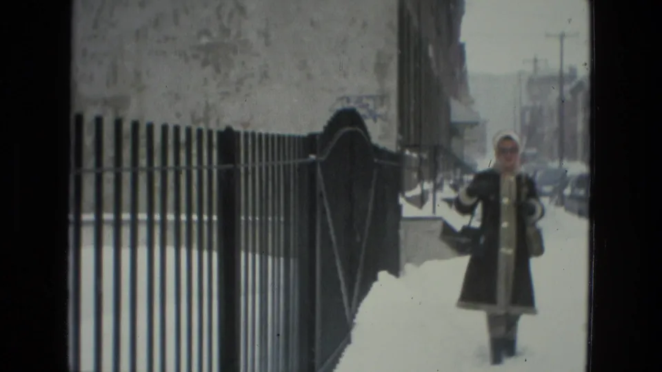 A bundled person walks along a snowy Drexel Hill sidewalk beside a metal fence, parked cars, and residential buildings.