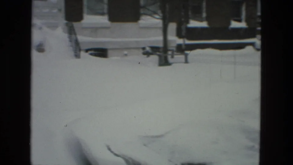 Snow-covered Drexel Hill street seen from indoors, with brick houses, bare tree branches, and parked cars softened by 8mm grain.