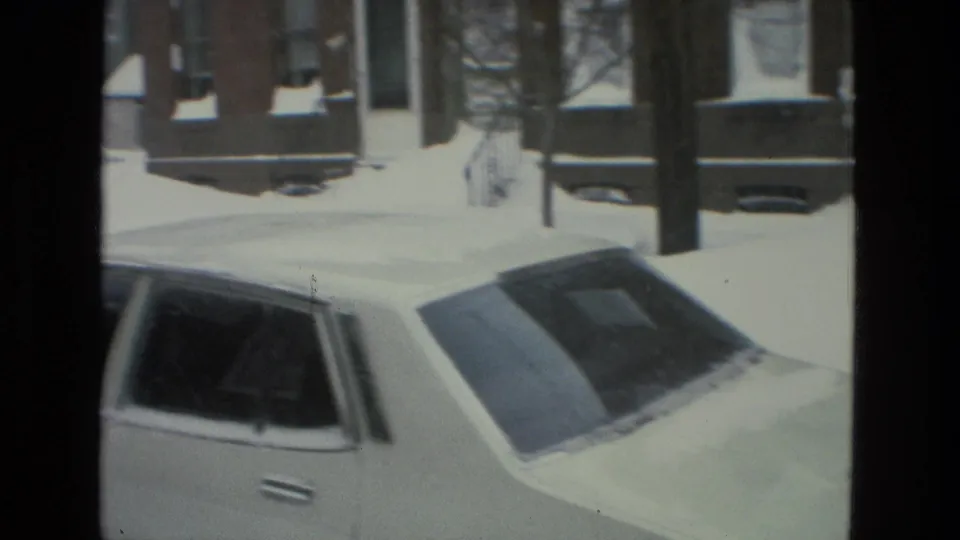 A parked car coated with snow sits along a Drexel Hill residential street, with brick row houses and bare trees in the background.