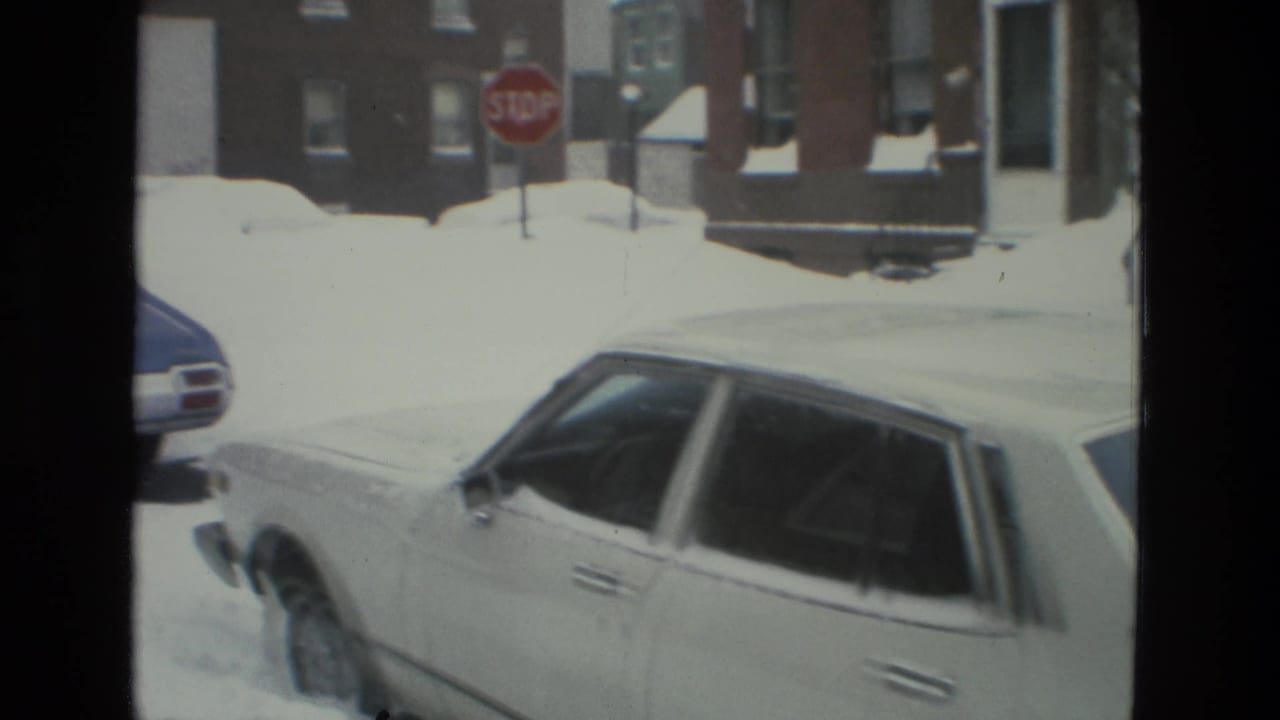 White sedan parked beside deep snowbanks with a stop sign and brick buildings beyond.