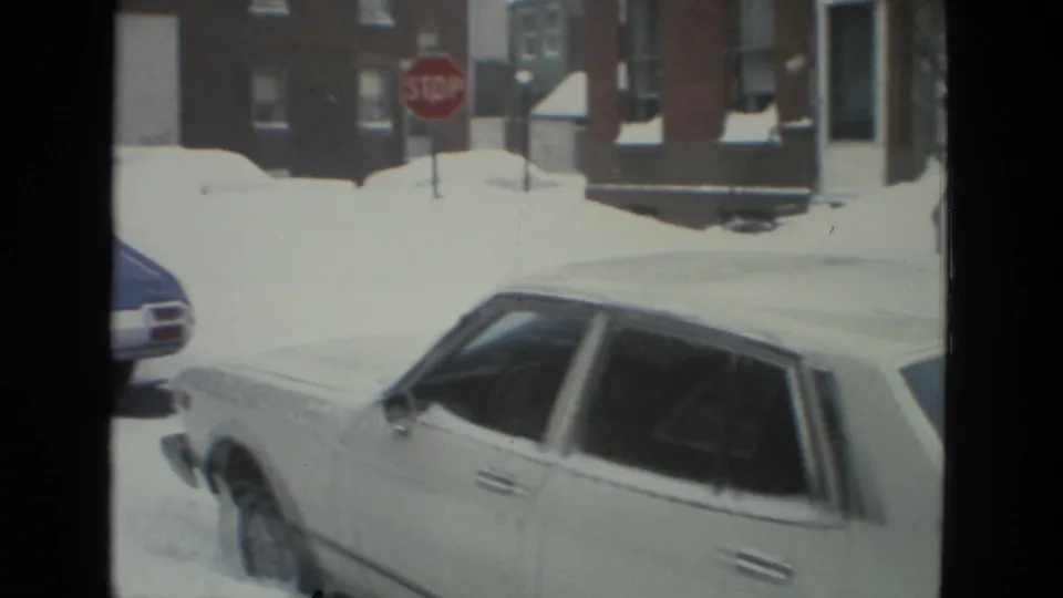 1978 Drexel Hill street view with a white sedan partly buried in snow near a red stop sign and brick residential buildings.