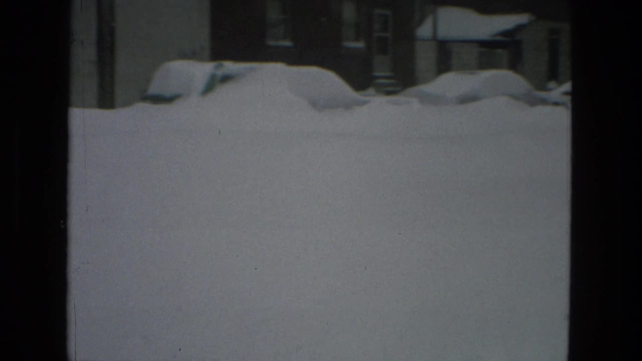 Large snowbank fills the foreground with parked cars and houses barely visible behind it.