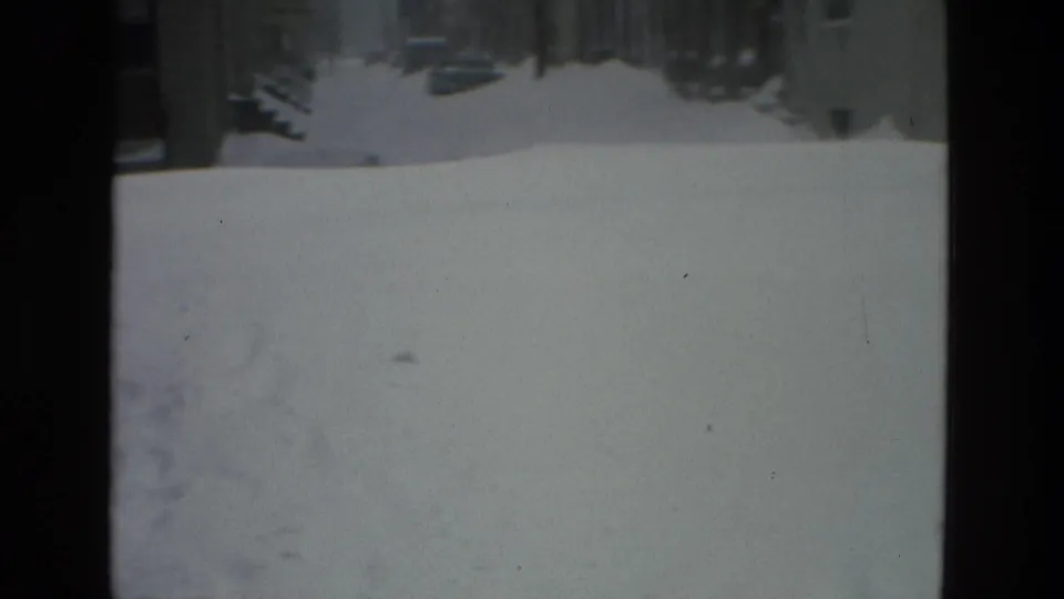 A snowy Drexel Hill street and sidewalk seen from indoors, with deep snowbanks and distant parked cars along the block.