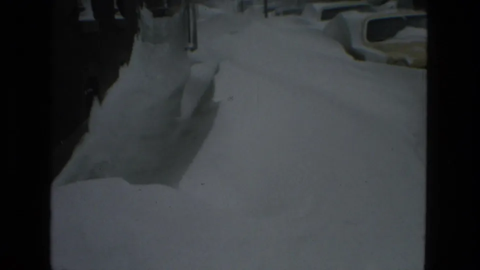 Snow piled along a narrow Drexel Hill walkway, with building walls, a downspout, and a parked car partly visible on the street.