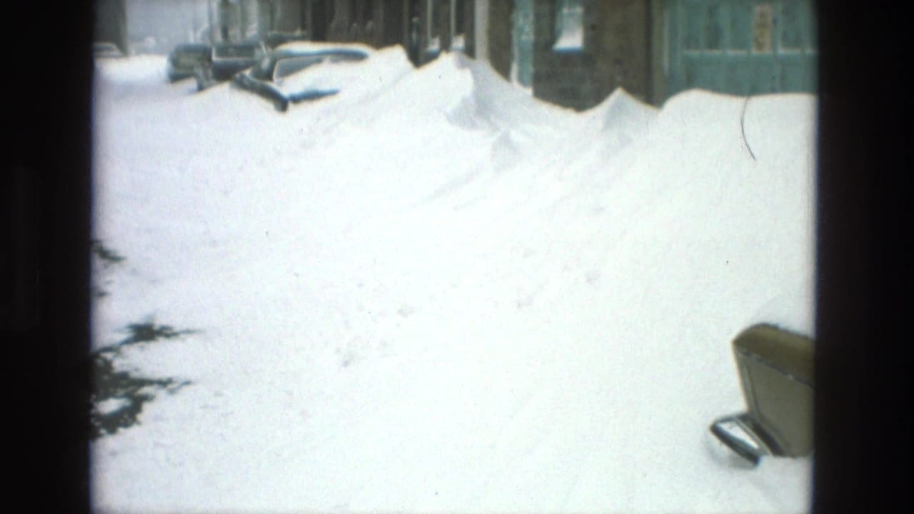 Bright snowy street with high drifts, parked cars, and buildings along the side.