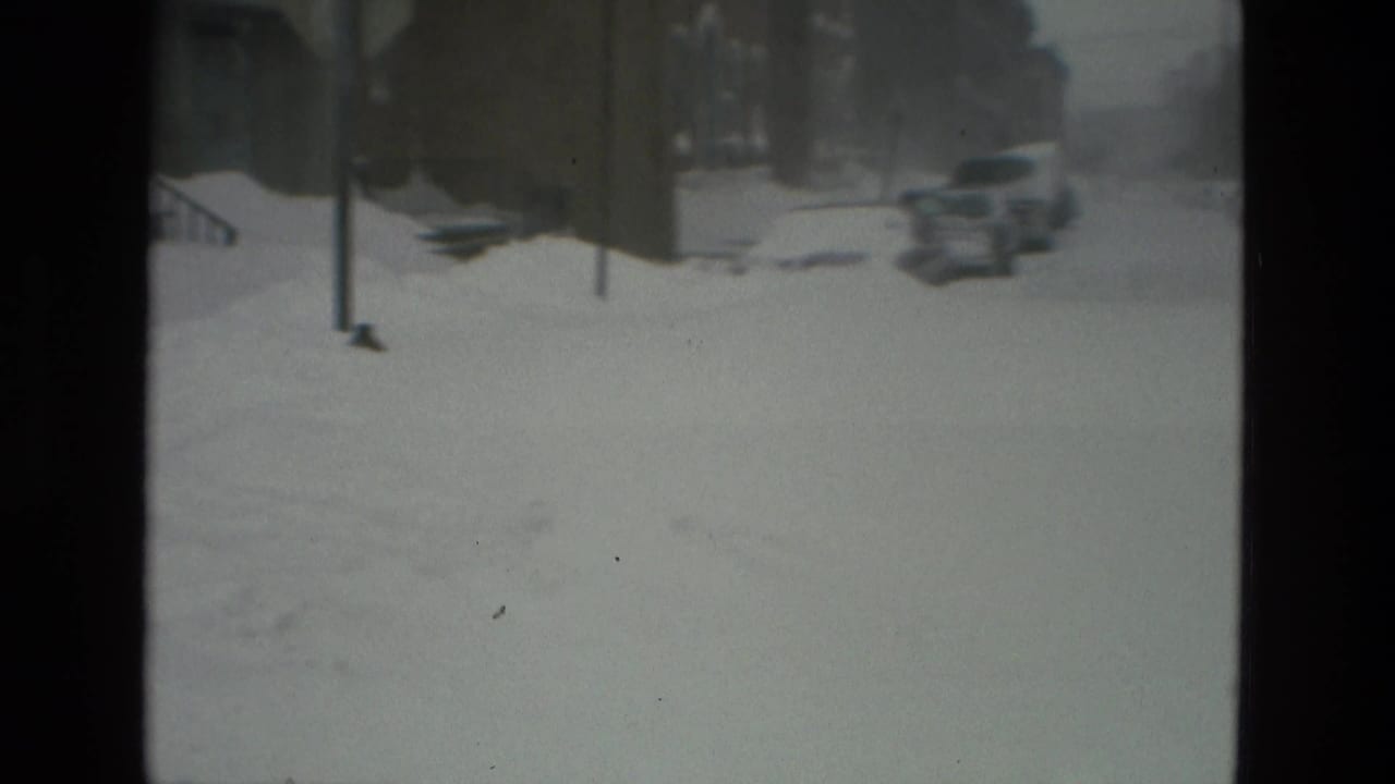 Snow-covered residential street with parked cars partly buried and buildings in soft focus.