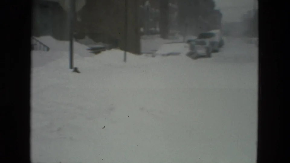 1978 Drexel Hill home-movie frame showing a snowbound street, buried cars, streetlamps, and residential buildings in muted 8mm color.