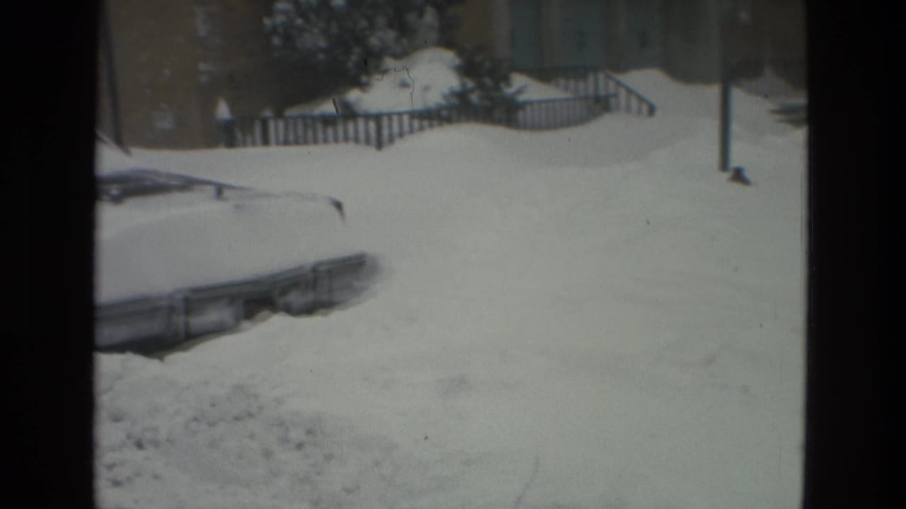 Parked car nearly covered by snow beside a residential building and railing.