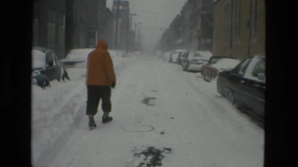 A distant person in an orange hooded coat walks along a snow-packed Drexel Hill street lined with parked cars in 1978 home-movie footage.