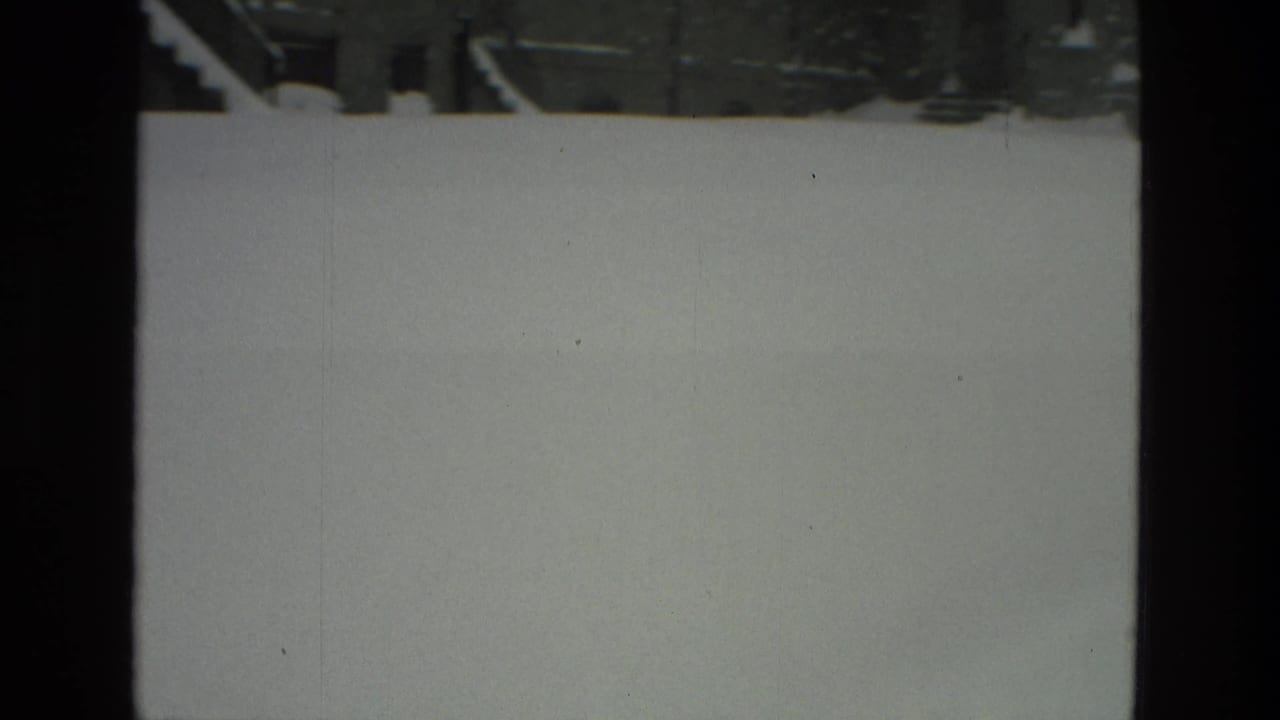 Snow blankets a Drexel Hill residential street in 1978, viewed through dark vertical window frames with parked cars faintly visible beyond the snow.