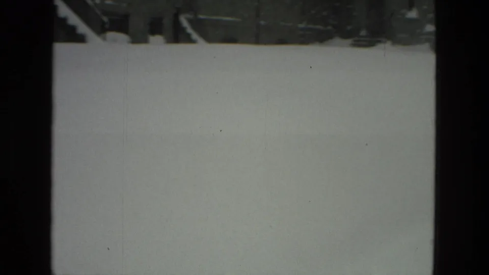 Snow blankets a Drexel Hill residential street in 1978, viewed through dark vertical window frames with parked cars faintly visible beyond the snow.