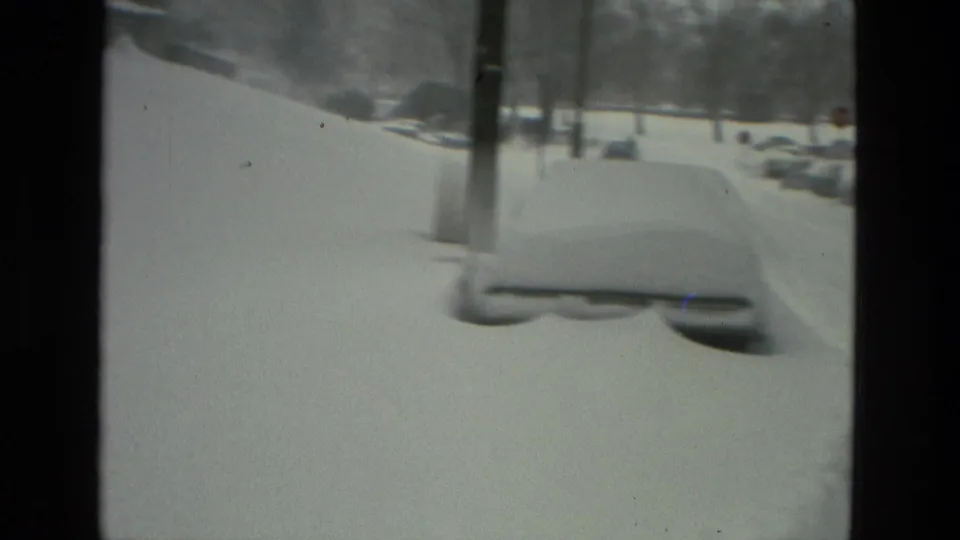 A snow-buried parked car sits near roadside poles on a Drexel Hill street during the 1978 winter storm, with the view softened by 8mm film grain.