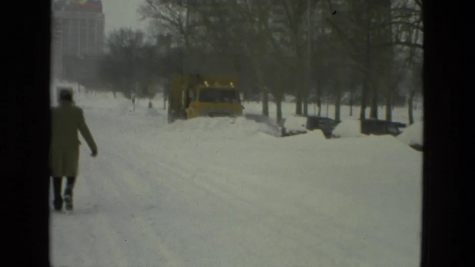 1978 Drexel Hill winter frame with a bundled person walking through snow as a yellow plow works ahead on the street.