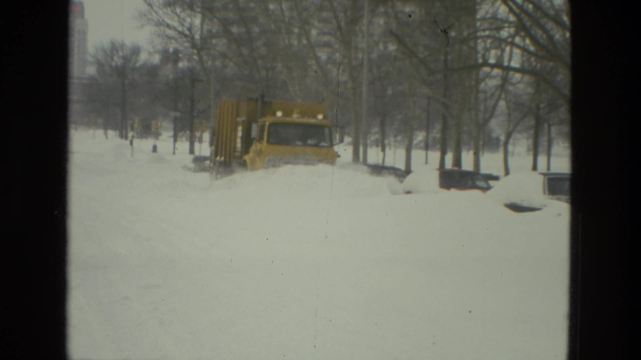 A yellow snowplow truck moves through deep snow on a tree-lined residential street in Drexel Hill, Pennsylvania, in 1978 home-movie footage.