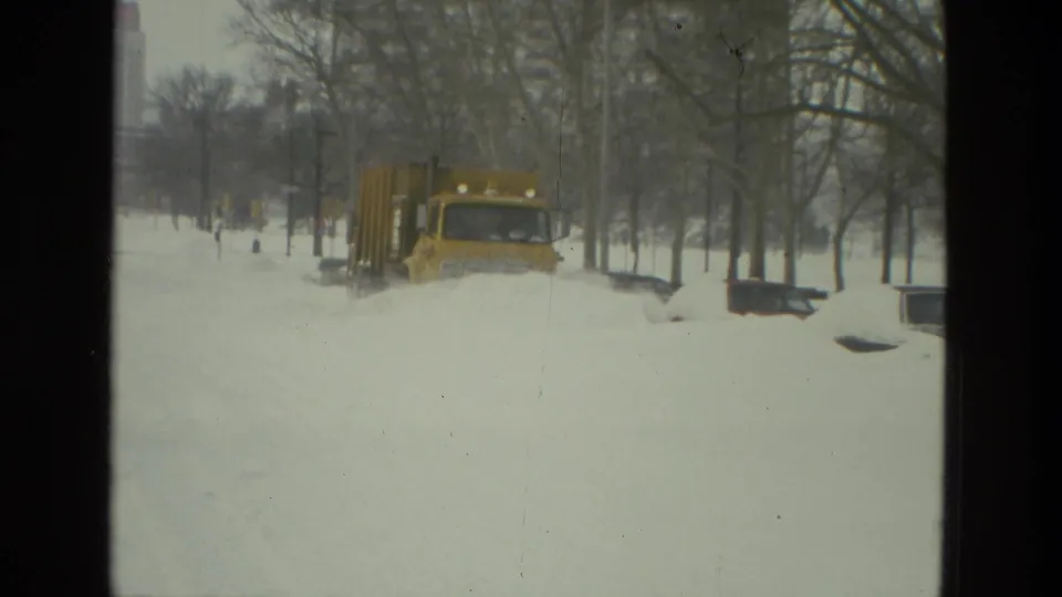 A yellow snowplow truck moves through deep snow on a tree-lined residential street in Drexel Hill, Pennsylvania, in 1978 home-movie footage.