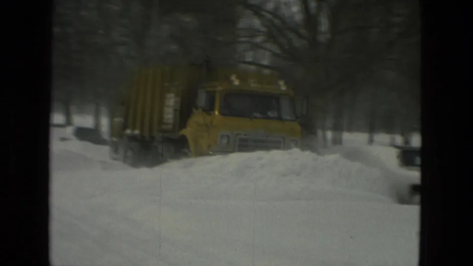 The yellow plow truck continues clearing a snow-covered Drexel Hill street, with bare trees and buried roadside cars visible through soft 8mm exposure.