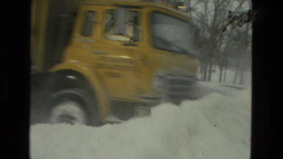 A close, slightly blurred 8mm frame shows the yellow snowplow truck passing near the camera as it pushes through a high snowbank in Drexel Hill.