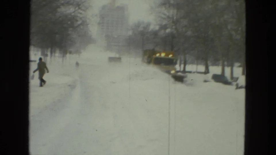 A distant pedestrian crosses or walks along the snowy Drexel Hill street as the yellow plow works near parked cars and bare winter trees in the background.