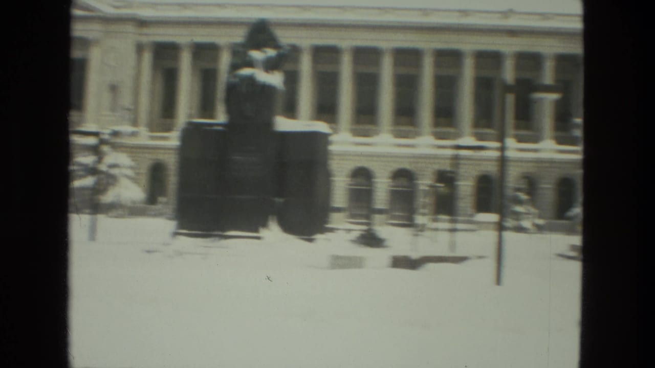 Snow-covered institutional building with columns and a dark statue in front.