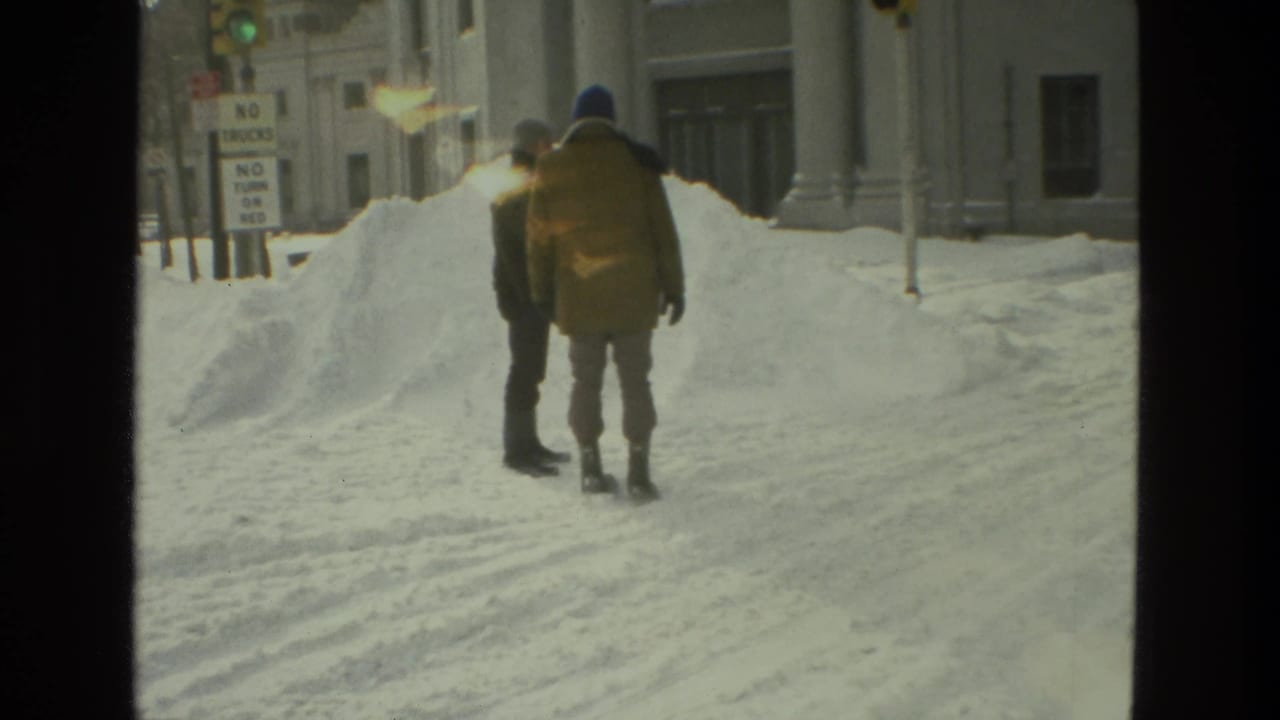 In 1978 Drexel Hill, Pennsylvania, two adults in winter coats walk away through deep snow beside urban buildings and traffic signs.
