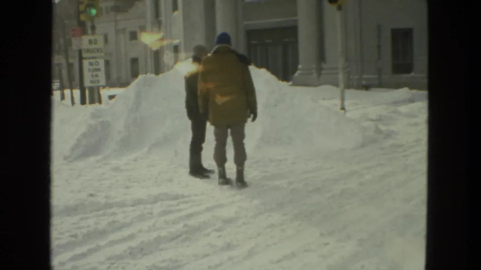 In 1978 Drexel Hill, Pennsylvania, two adults in winter coats walk away through deep snow beside urban buildings and traffic signs.