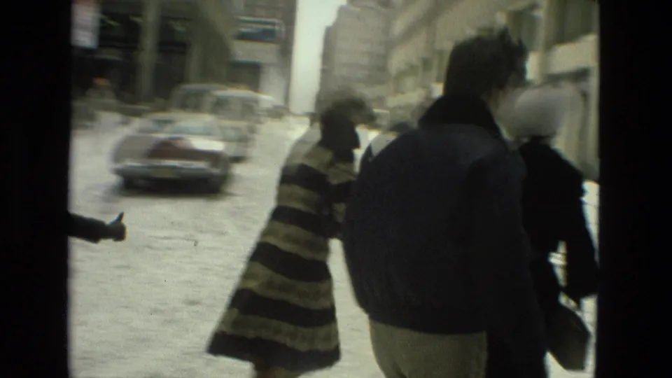 Adults in winter clothing gather near a snowy urban street while a car moves through the background in the 1978 footage.