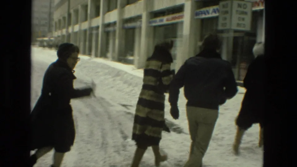 Bundled adults walk away on a snow-lined Drexel Hill sidewalk, with storefront windows and street signage visible behind them.