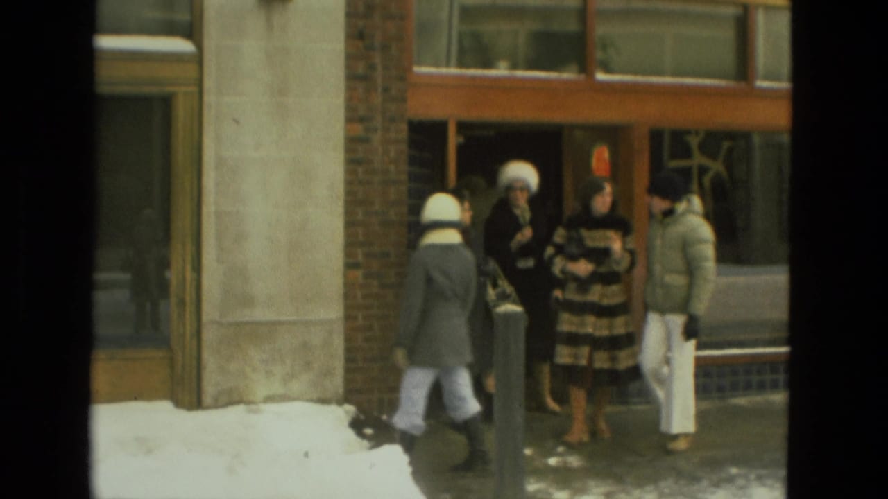 1978 Drexel Hill home-movie frame showing adults in winter coats gathered outside a brick-and-glass building with snow along the sidewalk.