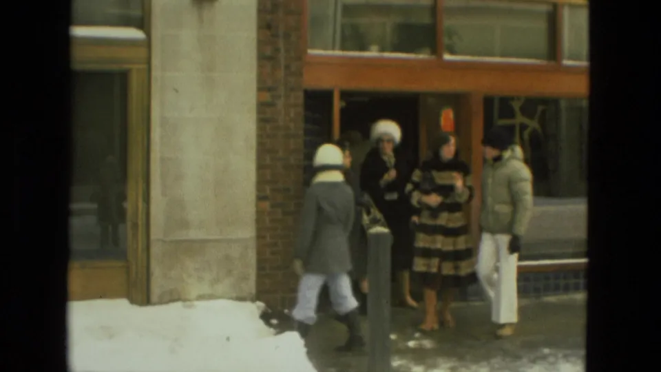 1978 Drexel Hill home-movie frame showing adults in winter coats gathered outside a brick-and-glass building with snow along the sidewalk.
