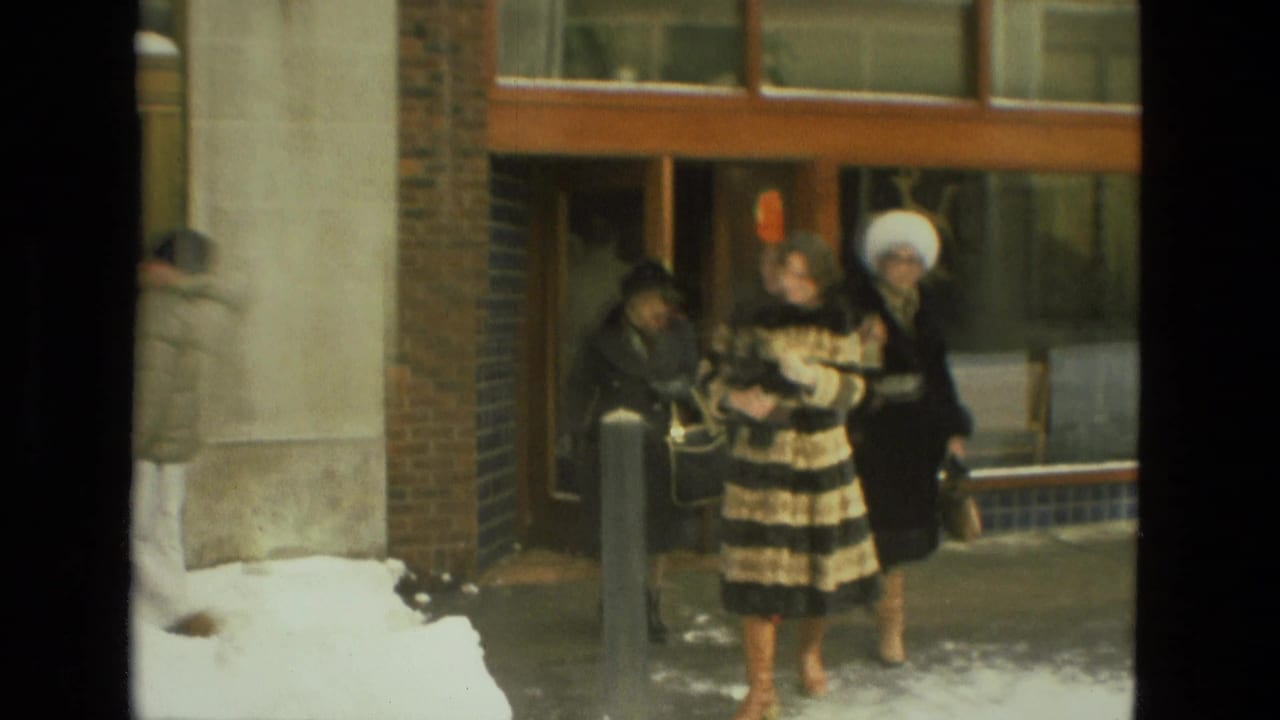 1978 Drexel Hill frame of two women, one in a striped fur-style coat and one in a white hat, walking outside a glass-fronted building.