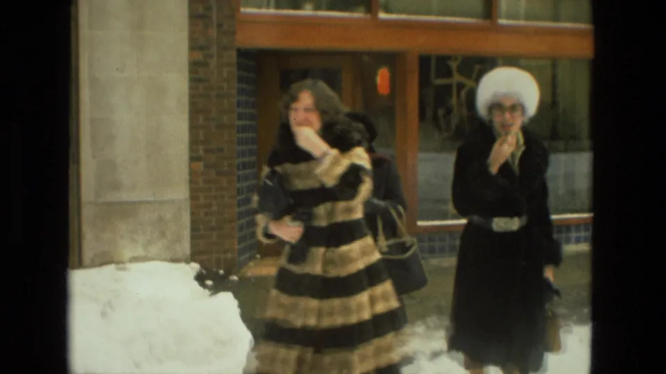1978 Drexel Hill home-movie frame with two women approaching the camera outside a brick-and-glass building in winter clothing.