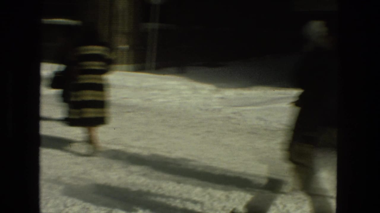 Blurred pedestrians walk across a snowy street beside dark buildings.