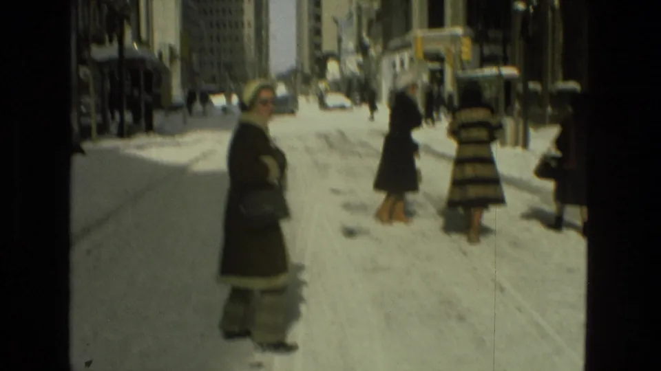 1978 Drexel Hill home-movie frame showing women in winter coats on a snowy street lined with buildings and parked cars.