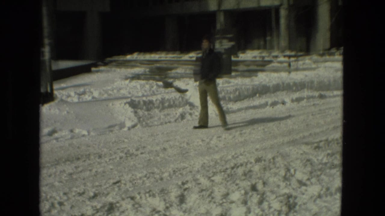 A person walks across a snow-covered open area near large buildings.