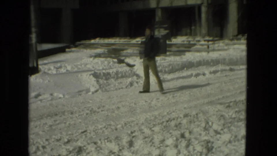 A bundled pedestrian crosses a snowy paved area near modern buildings in this 1978 Drexel Hill home-movie frame.