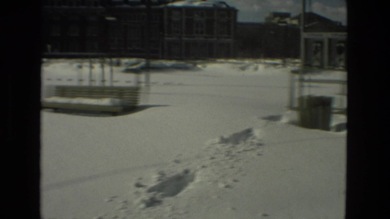 Snow-covered public square with benches, footprints, a trash can, and buildings in the distance.