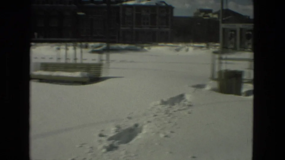 1978 Drexel Hill home-movie frame showing snow-covered ground, benches, footprints, a trash can, and distant buildings.
