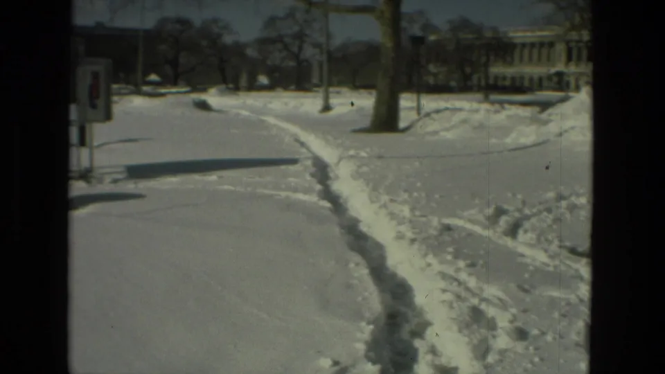 1978 Drexel Hill frame with a curved snowy footpath, poles, lampposts, bare trees, and institutional-looking buildings beyond.