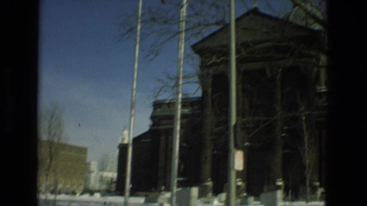 Snowy street view of a dark masonry building with columns, flagpoles, and bare trees.