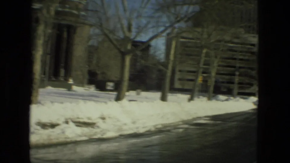 Moving-vehicle 8mm view of a columned building entrance, roadside snow, bare trees, and a neighboring structure in Drexel Hill, Pennsylvania.