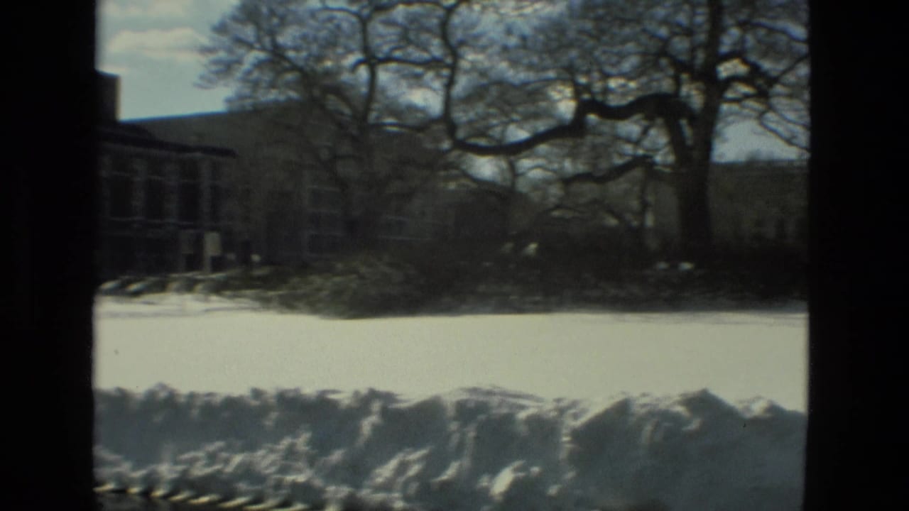 Snow-covered grounds with bare trees and distant institutional buildings behind dark frame edges.