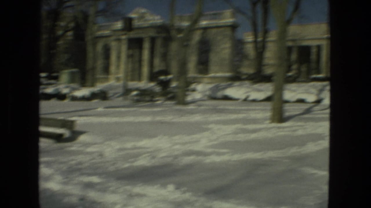 Snow-covered park street with benches, bare trees, and a columned stone building in the background.