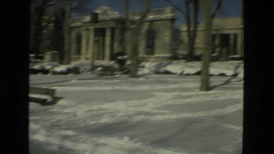1978 Drexel Hill winter view with deep snow, bare trees, benches, parked cars, and a columned stone building beyond the road.
